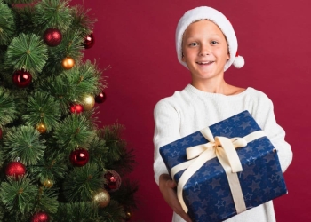 Criança com câncer segurando um presente ao lado da árvore de Natal, para representar a campanha da cartinha de Natal.