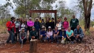 Grupo de voluntários reunido na Floresta Nacional do DF, em uma área de solo avermelhado e cercada por árvores, participando de um mutirão de replantio para restaurar a vegetação afetada pelas queimadas. Todos estão equipados para a atividade e posam juntos e sorridentes.