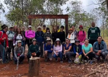 Grupo de voluntários reunido na Floresta Nacional do DF, em uma área de solo avermelhado e cercada por árvores, participando de um mutirão de replantio para restaurar a vegetação afetada pelas queimadas. Todos estão equipados para a atividade e posam juntos e sorridentes.