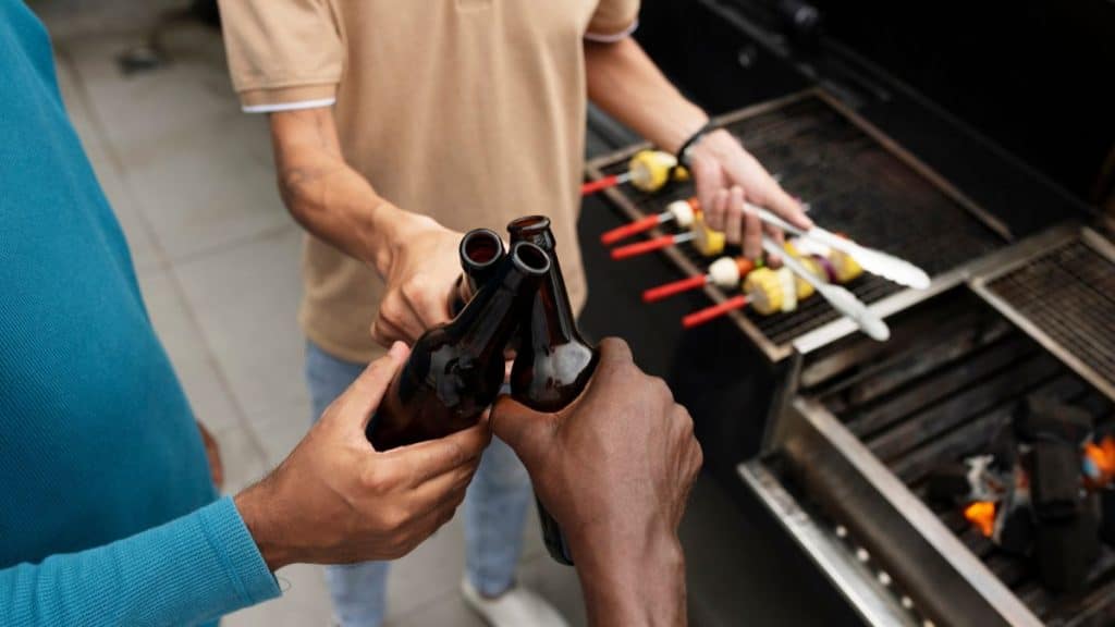 Três pessoas brindando com garrafas de bebida enquanto preparam espetinhos na churrasqueira.