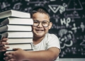 Menino sorrindo com pilha de livros em frente a quadro com fórmulas matemáticas.