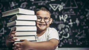 Menino sorrindo com pilha de livros em frente a quadro com fórmulas matemáticas.