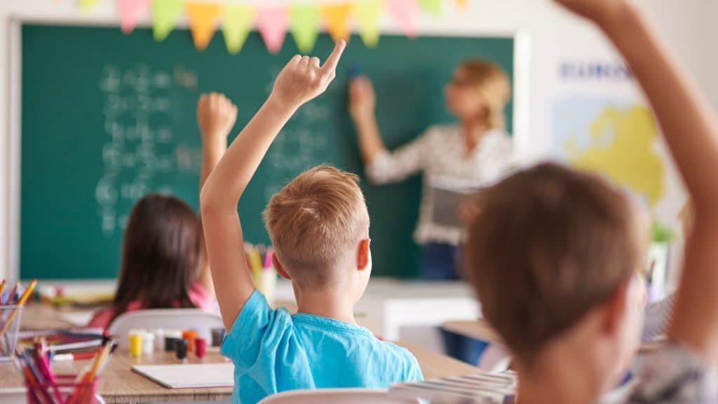 Crianças em sala de aula levantando as mãos enquanto uma professora escreve no quadro.