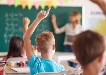 Crianças em sala de aula levantando as mãos enquanto uma professora escreve no quadro.