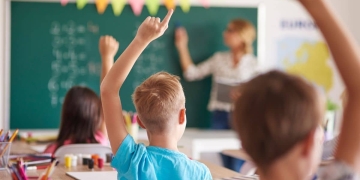 Crianças em sala de aula levantando as mãos enquanto uma professora escreve no quadro.