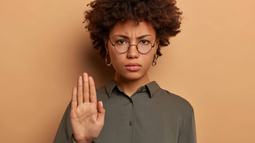 Uma mulher de expressão séria e decidida levanta a mão com a palma virada para frente, em um gesto de "pare" ou recusa. Ela usa óculos redondos e veste uma camisa verde. Ao fundo, um tom de marrom claro reforça o foco em sua expressão determinada.