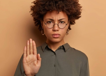 Uma mulher de expressão séria e decidida levanta a mão com a palma virada para frente, em um gesto de "pare" ou recusa. Ela usa óculos redondos e veste uma camisa verde. Ao fundo, um tom de marrom claro reforça o foco em sua expressão determinada.