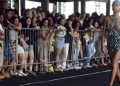 Modelo caminha em uma passarela ao ar livre durante o Desfile Beleza Negra, vestindo uma roupa estilizada com cores vibrantes. Ao fundo, uma plateia diversa observa o evento, destacando o impacto cultural e social da iniciativa.