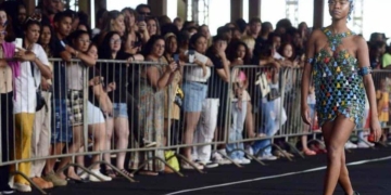 Modelo caminha em uma passarela ao ar livre durante o Desfile Beleza Negra, vestindo uma roupa estilizada com cores vibrantes. Ao fundo, uma plateia diversa observa o evento, destacando o impacto cultural e social da iniciativa.