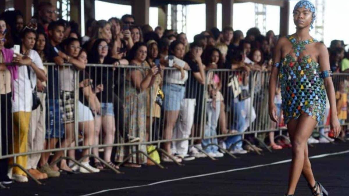 Modelo caminha em uma passarela ao ar livre durante o Desfile Beleza Negra, vestindo uma roupa estilizada com cores vibrantes. Ao fundo, uma plateia diversa observa o evento, destacando o impacto cultural e social da iniciativa.