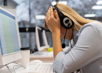 Funcionária com fone de ouvido apoia a cabeça nas mãos em frente ao computador, aparentando cansaço.