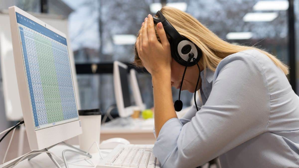 Funcionária com fone de ouvido apoia a cabeça nas mãos em frente ao computador, aparentando cansaço.