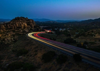 Rodovia iluminada com trilhas de luz dos faróis de veículos em movimento, situada em um cenário montanhoso ao anoitecer.