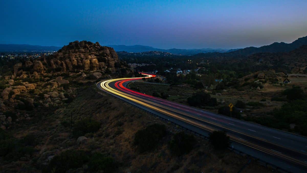 Rodovia iluminada com trilhas de luz dos faróis de veículos em movimento, situada em um cenário montanhoso ao anoitecer.