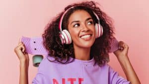 Mulher sorridente com cabelos cacheados usando fones de ouvido rosa e branco, segurando um skate lilás em frente a um fundo rosa.