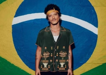 Cantor Bruno Mars parado na frente de uma bandeira do Brasil pintada na parede.