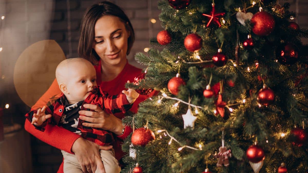 Mulher segurando bebê ao lado da árvore de natal.