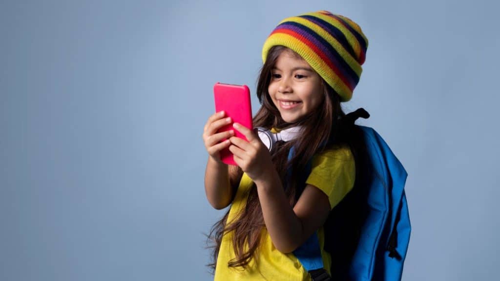 Uma criança sorridente usa um celular rosa, vestindo uma touca colorida e mochila azul, em frente a um fundo azul.