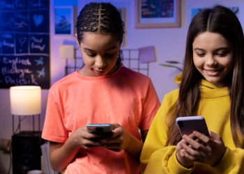 uas adolescentes utilizando celulares em ambiente escolar, com quadro de tarefas ao fundo.