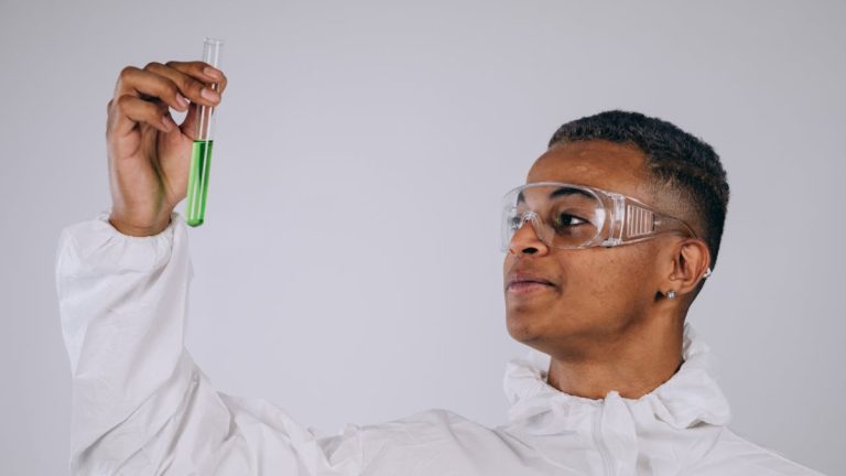 Jovem cientista segurando um tubo de ensaio com líquido verde, usando óculos de proteção e jaleco branco em ambiente de laboratório.