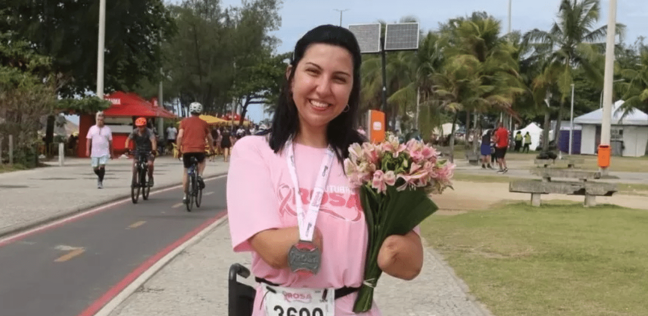 Mulher sorri após corrida, com flores na mão e medalha no peito, representando superação após meningite.