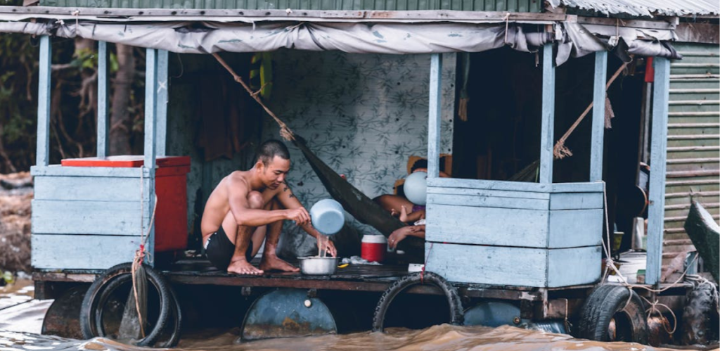 Homem em casa flutuante cozinha enquanto a água da enchente invade a área ao redor. No fundo, outra pessoa descansa em uma rede, adaptando-se às dificuldades causadas pelo alagamento.