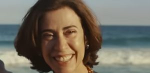 Mulher sorridente em frente ao mar, com cabelos curtos e colar de contas, capturada em um momento de felicidade à beira da praia