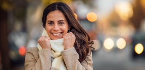 Mulher sorridente vestindo um casaco bege e um cachecol, segurando a gola para se proteger do frio, com luzes desfocadas ao fundo em um ambiente urbano