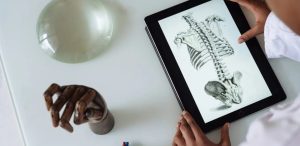 Uma pessoa vestindo um jaleco branco interage com um tablet que exibe uma ilustração anatômica da coluna vertebral e costelas humanas. Sobre a mesa branca, há uma mão de madeira articulada, uma cúpula translúcida