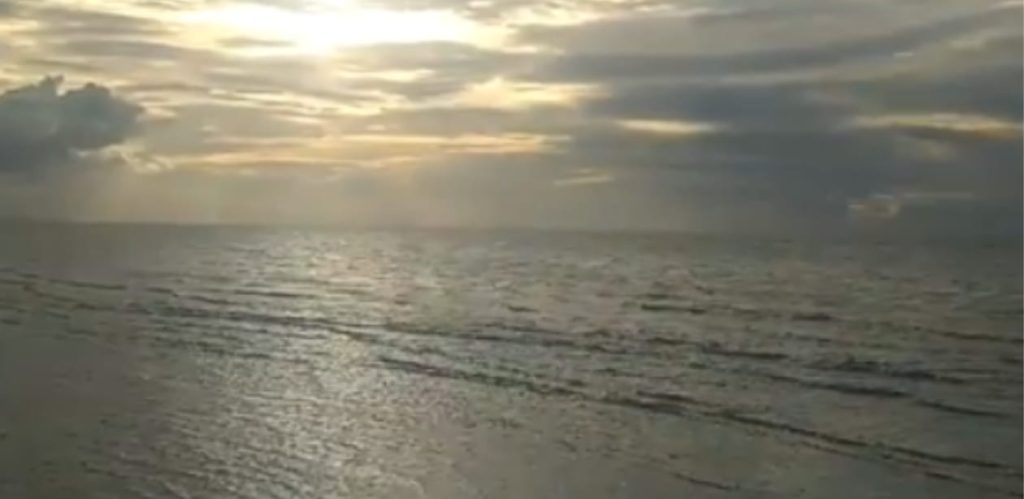 Vista panorâmica da Praia de Arpoeiras ao entardecer, com o mar calmo refletindo a luz dourada do sol entre nuvens no horizonte.