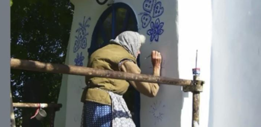 A imagem mostra uma mulher idosa pintando delicados desenhos florais e de borboletas em uma parede branca. Ela está vestindo um lenço na cabeça, um colete marrom sobre uma blusa de mangas compridas e uma saia azul com estampa de bolinhas. A mulher parece estar em um andaime improvisado, trabalhando com um pincel e um pequeno recipiente de tinta azul. Ao fundo, há uma porta azul arredondada e algumas árvores que fornecem sombra. A cena transmite um ambiente rústico e artesanal, possivelmente representando uma tradição cultural de decoração de casas.