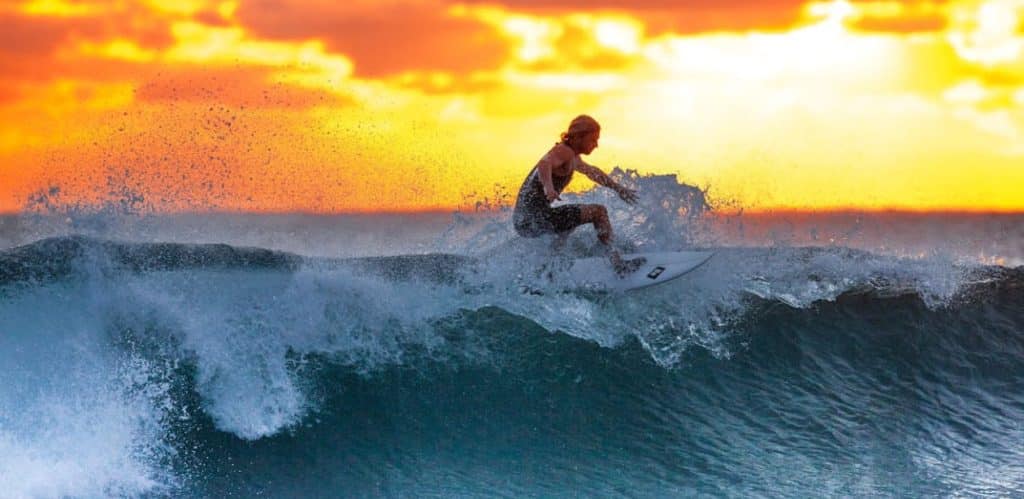 Surfista manobrando no topo de uma onda ao pôr do sol, com o céu alaranjado ao fundo e a água espirrando com o impacto da prancha. A cena captura a energia e a liberdade do surfe