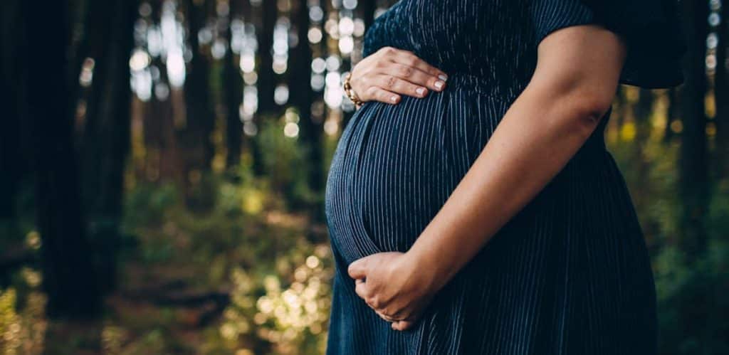 Mulher grávida vestindo um vestido azul escuro segura delicadamente sua barriga enquanto caminha em um ambiente natural com árvores ao fundo