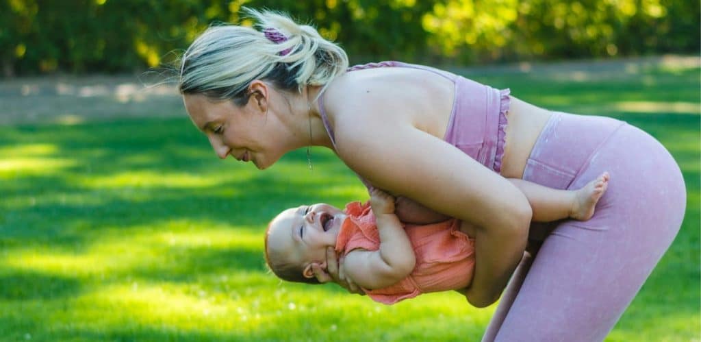 Mãe praticando atividade física ao ar livre enquanto segura seu bebê no colo, ambos sorrindo. Exercício no pós-parto melhora o bem-estar.