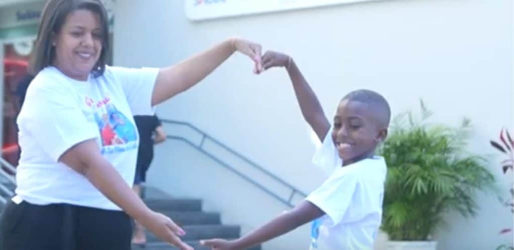 Mãe e filho comemoram a vitória contra a leucemia, formando um coração com as mãos em frente ao hospital. Ambos sorriem, simbolizando superação e esperança