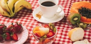 A imagem mostra um café da manhã saudável e vibrante, com uma variedade de frutas frescas, como morangos, banana, kiwi e mamão. Ao lado, uma xícara de café, um prato com frutas em cubos e uma fatia de pão completam a refeição. A mesa está disposta sobre uma toalha xadrez vermelha e branca, criando um ambiente acolhedor e saboroso para começar o dia com energia. A variedade de frutas adiciona cores e nutrientes importantes, tornando o café da manhã uma refeição rica e equilibrada.