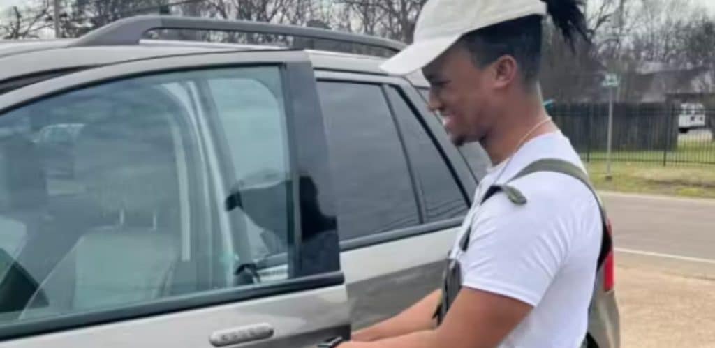 A imagem mostra um jovem sorridente diante de um carro novo, prestes a entrar nele. Ele parece emocionado e surpreso com a novidade, demonstrando uma expressão de felicidade ao receber o presente inesperado da comunidade