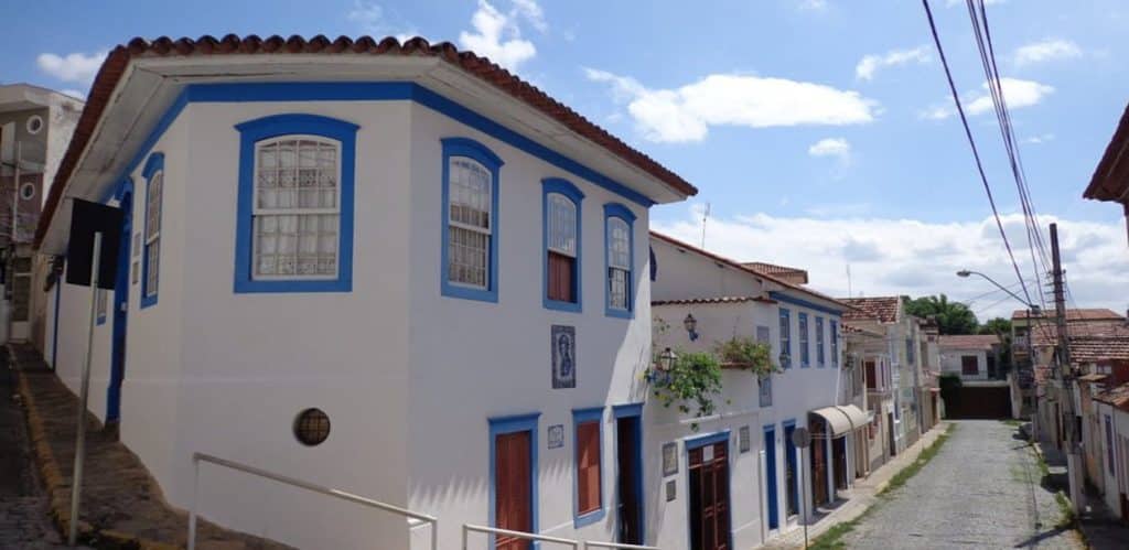 A imagem mostra uma rua tranquila com casas de arquitetura colonial, destacando-se uma residência de fachada branca com detalhes em azul, típica das cidades históricas brasileiras. As janelas possuem molduras em azul, que contrastam com a parede branca. No cenário, é possível ver um céu claro, com poucas nuvens, e ruas calmas, pavimentadas com pedras. A cena transmite uma sensação de paz e simplicidade, comum em muitas cidades do interior, que preservam sua história e charme.