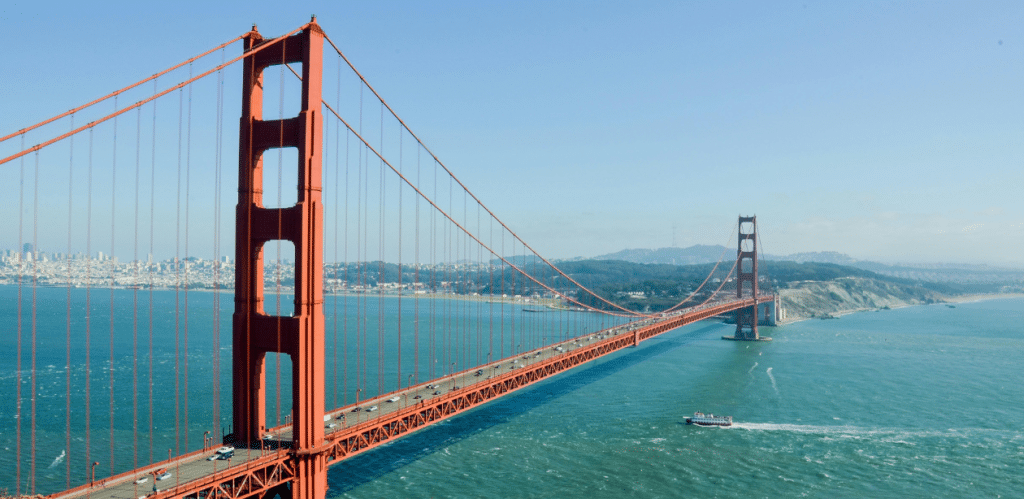 A imagem mostra a famosa Ponte Golden Gate, localizada na Califórnia, EUA. A ponte, com sua estrutura vermelha icônica, se estende sobre as águas do oceano, conectando a cidade ao condado de Marin. Ao fundo, é possível ver a linha do horizonte de San Francisco, com suas colinas e a costa ao longe. A cena é clara e ensolarada, com um barco navegando nas águas calmas, criando uma vista panorâmica impressionante e destacando a grandiosidade da ponte.