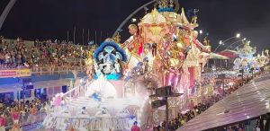 A imagem captura um desfile de carnaval à noite, com um carro alegórico grandioso, passando pelo sambódromo. O carro tem uma temática dourada e rosa, adornado com esculturas brilhantes de figuras femininas segurando cálices dourados, penas azuis exuberantes. Há uma grande multidão assistindo das arquibancadas, com muitas luzes iluminando o evento. brilho e celebração.