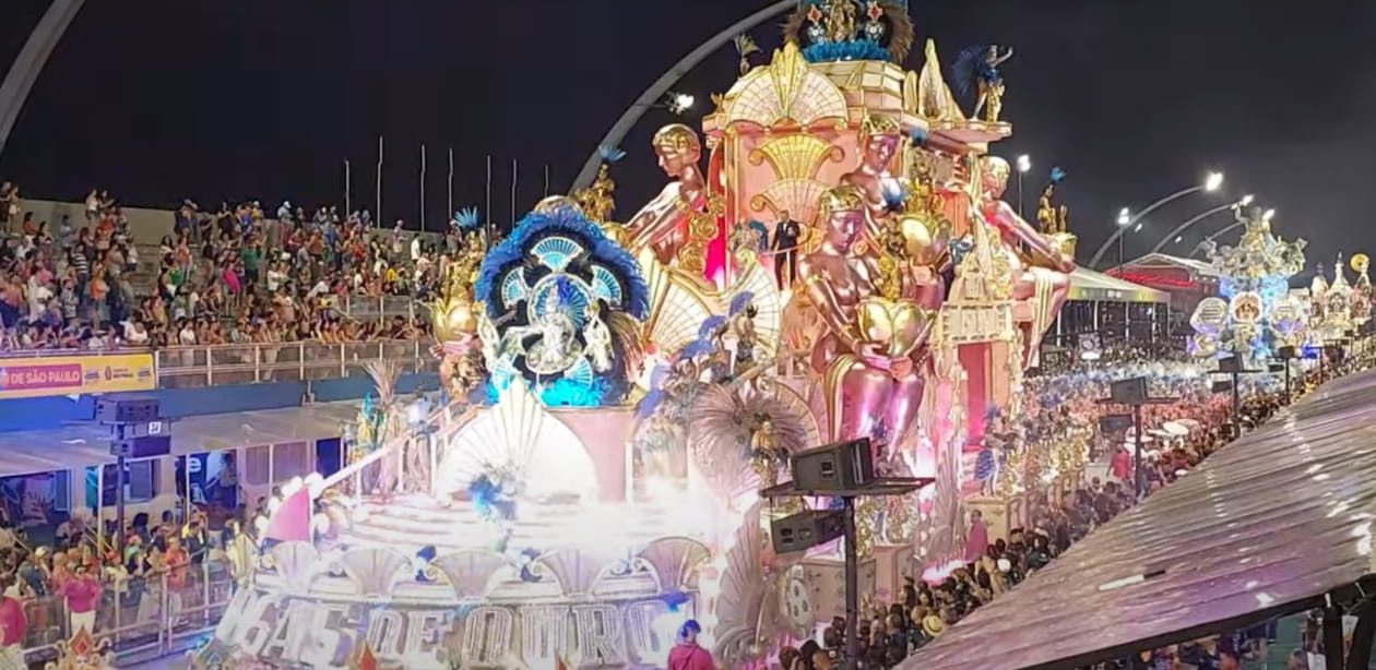 A imagem captura um desfile de carnaval à noite, com um carro alegórico grandioso, passando pelo sambódromo. O carro tem uma temática dourada e rosa, adornado com esculturas brilhantes de figuras femininas segurando cálices dourados, penas azuis exuberantes. Há uma grande multidão assistindo das arquibancadas, com muitas luzes iluminando o evento. brilho e celebração.