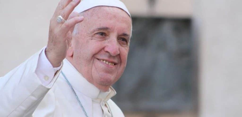 Papa Francisco aparece sorridente, vestindo sua tradicional veste branca e acenando com a mão. O pontífice transmite serenidade e proximidade, enquanto é registrado em um ambiente externo com fundo levemente desfocado. A imagem simboliza sua conexão com os fiéis e sua presença marcante como líder da Igreja Católica.