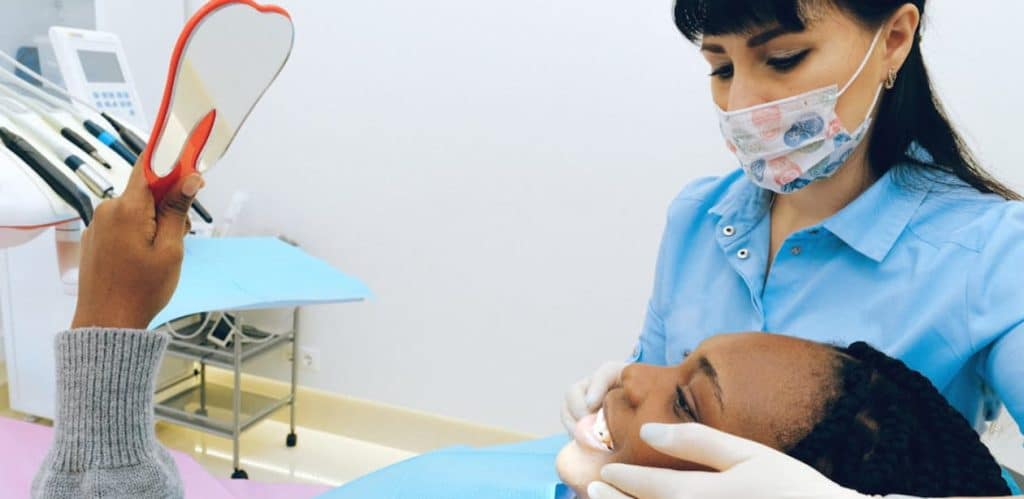 Uma paciente negra está deitada na cadeira de um consultório odontológico, segurando um espelho em formato de dente para visualizar seu sorriso. A dentista, vestindo um uniforme azul e máscara facial estampada, examina a boca da paciente com luvas e instrumentos odontológicos. O ambiente é bem iluminado e organizado, com equipamentos odontológicos ao fundo.