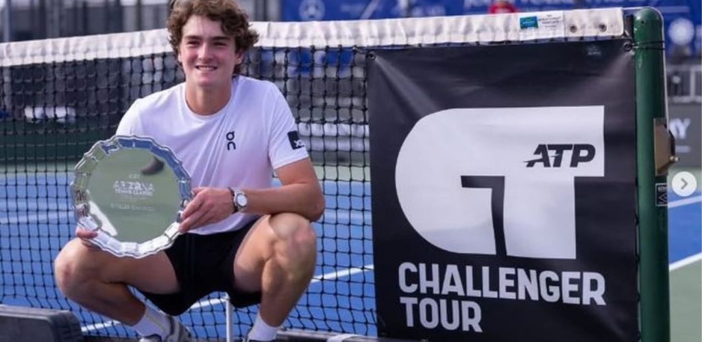 Tenista brasileiro João Fonseca posa sorridente ao lado da rede segurando o troféu do Challenger Tour, após vencer torneio em Phoenix.