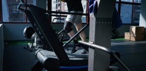 Pessoa treinando em esteira em academia, usando joelheira esportiva. Ambiente iluminado com janelas grandes e equipamentos de academia.