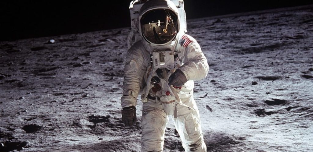 Astronauta caminha na superfície lunar vestindo traje espacial completo. O reflexo do capacete captura a paisagem da Lua, destacando a importância das missões espaciais para a exploração do universo.