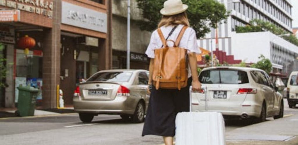 Mulher viajante caminha sozinha em uma cidade, carregando uma mala branca e mochila, simbolizando o turismo solo feminino