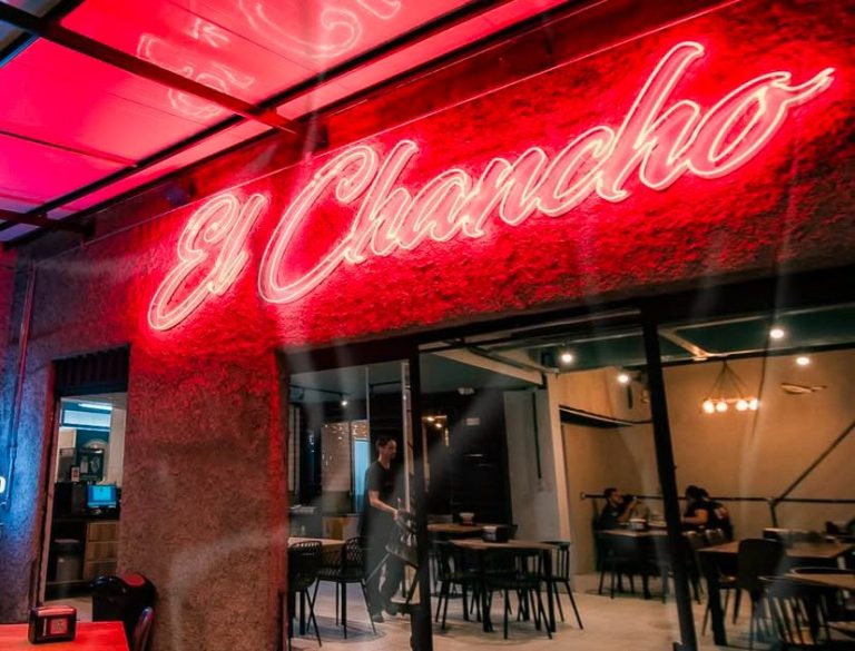 Fachada de um restaurante chamado 'El Chancho' com um letreiro de neon vermelho brilhante. A entrada tem uma parede de textura rústica e uma grande abertura de vidro que revela o interior do local. Dentro, algumas mesas e cadeiras estão organizadas, com clientes sentados e um funcionário em pé arrumando o espaço. A iluminação aconchegante e o teto parcialmente coberto contribuem para um ambiente convidativo