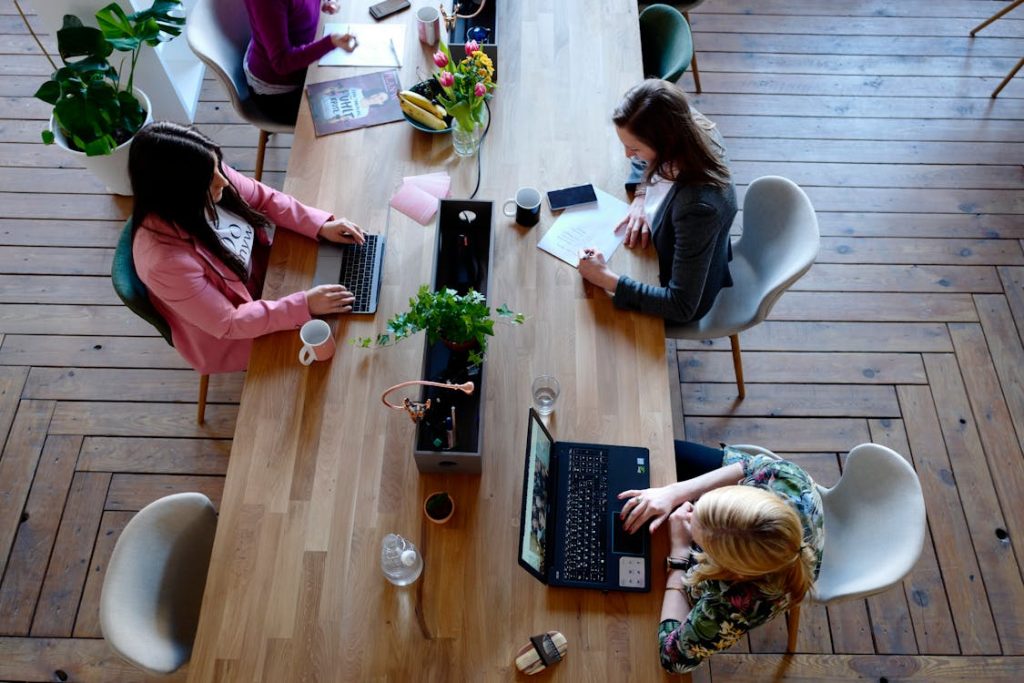 Três mulheres trabalhando em um escritório colaborativo moderno, sentadas ao redor de uma mesa de madeira, utilizando laptops e tomando notas