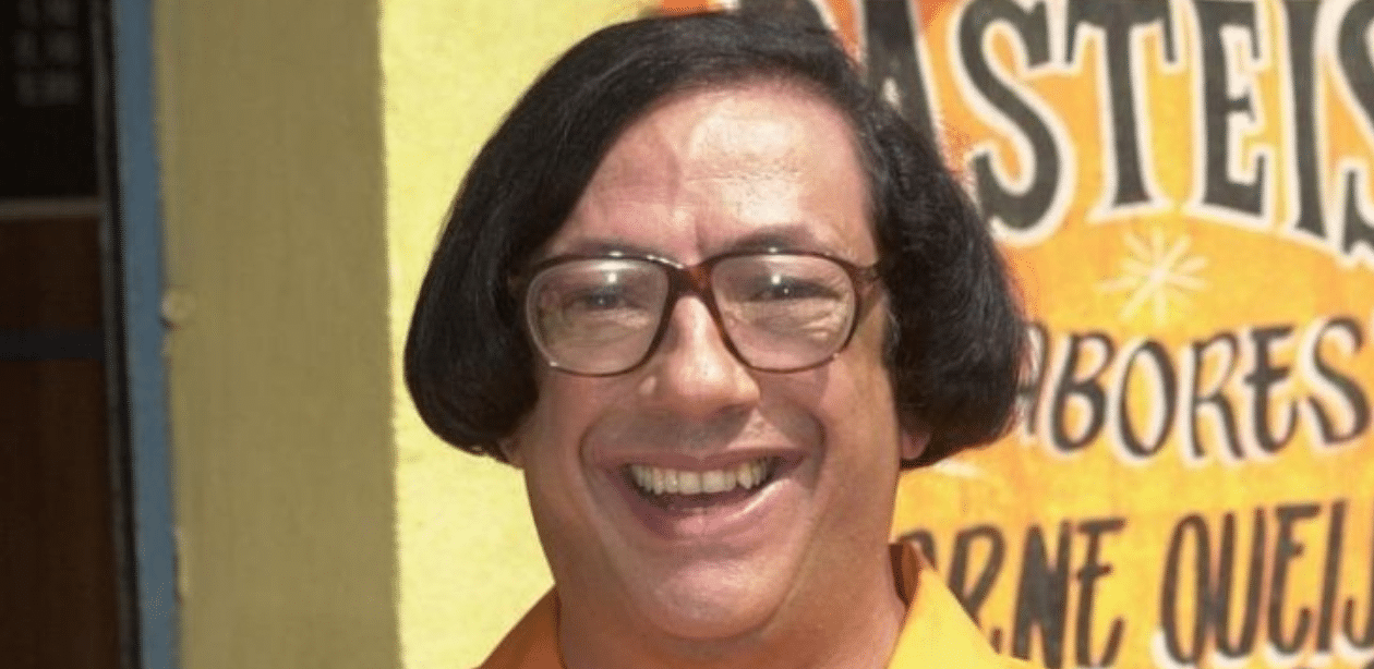 Marcos Oliveira caracterizado como Beiçola em cena da série "A Grande Família", sorrindo em frente a uma parede com letreiro de pastelaria.